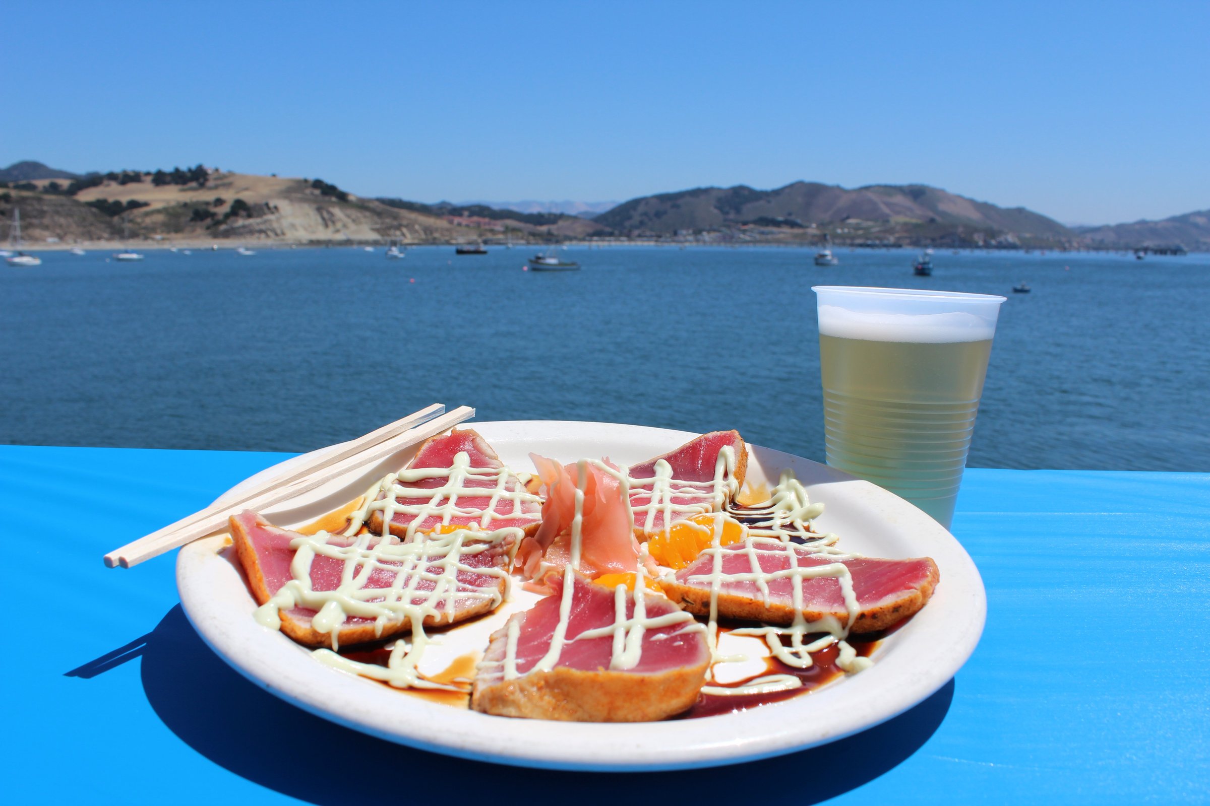 Seared ahi tuna with wasabi cream and a cold beer on a blue table at Port San Luis, with boats moored in San Luis Bay behind